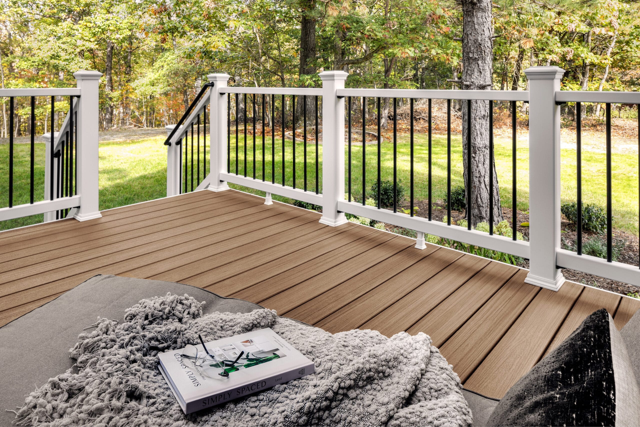 Backyard deck built with Trex Enhance Golden Hour decking, featuring warm golden-brown boards, white railings, and a cozy lounge area with a blanket and open book, highlighting the inviting, natural wood-grain texture.