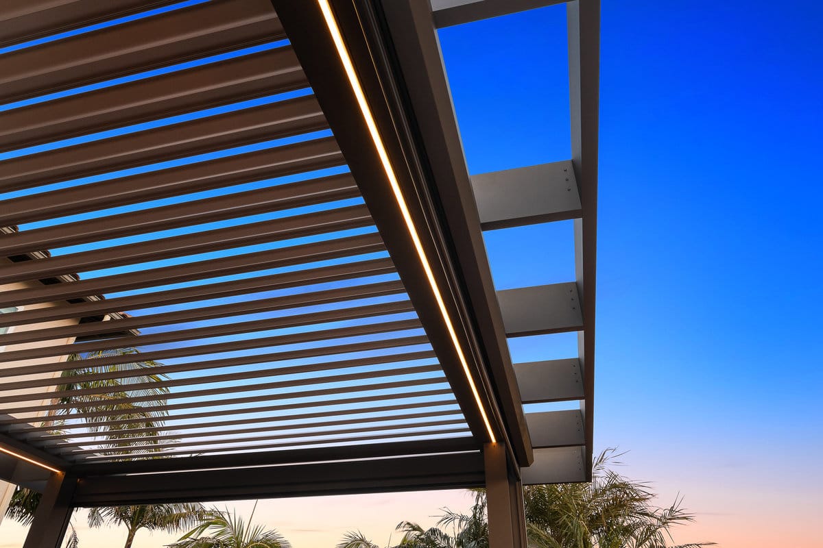 Motorized louvered pergola with integrated lighting and open slats revealing a clear blue sky.